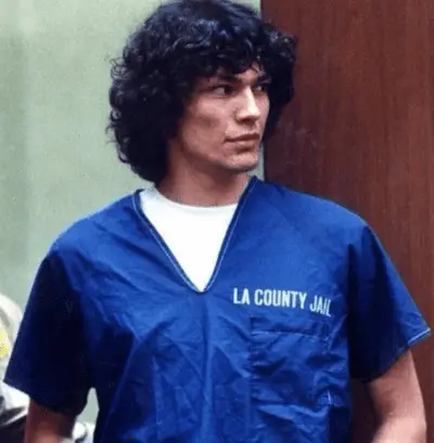 A picture of Richard Ramirez. He is wearing a blue LA County prisoner jumpsuit. He is wearing a white t shirt underneath. His dark hair is a bit longer. 