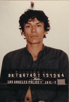 Richard Ramirez Police Photo. Richard has a dark blue shirt with his stomach exposed. His hair is black and curly. The button of his shirt is unbuttoned.