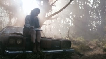 A young woman sits on top of an abandoned car smoking and drawing in a notebook.