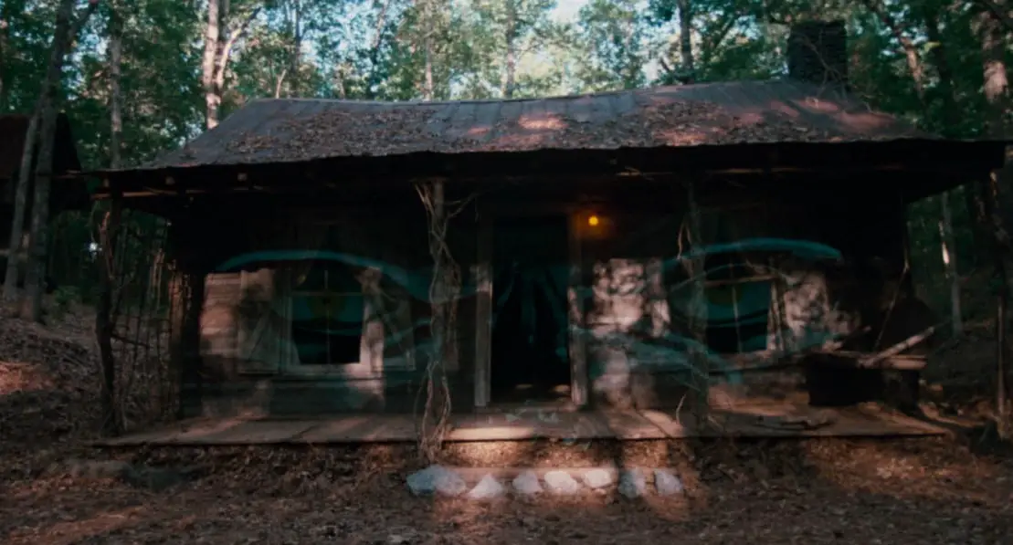 A shot of an old, abandoned looking cabin with a pair of angry looking eyes layered over the image.