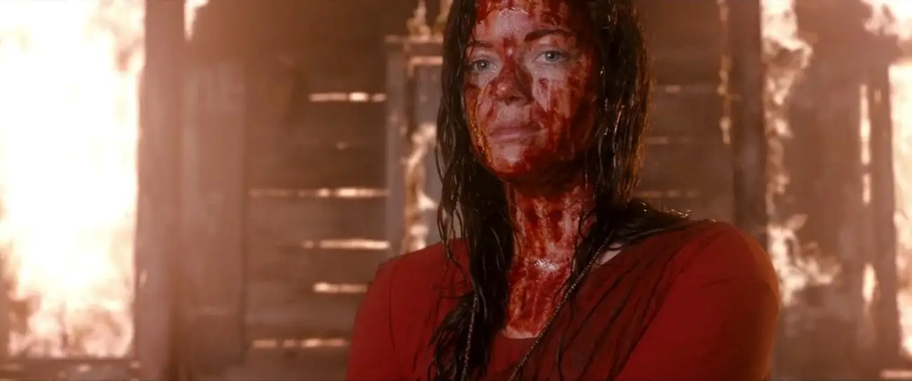 A young woman with blood on her face stands in front of a burning building looking out into the distance.