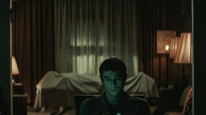 Yakov sits looking at his phone with his back turned to the outline of a body on a gurney in the room behind him