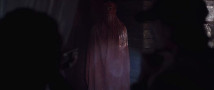 Ivy points a light and a shotgun at a figure dressed in a red hooded cloak in the corner of a cabin