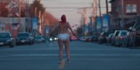 A woman walks in the center of the street away from the camera her bare back is seen and she is only wearing underwear. Her head is covered in a red wax substance and her arms and legs have duct tape around them.