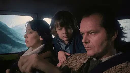 The Torrances in the car on the way to The Overlook; Jack is at the wheel, Wendy is beside him in the passenger seat, looking straight ahead; Danny has pulled himself up from the back seat and rests his arms on the backs of his parents' seats; he is looking at his father, whose eyes are pointed upward, suggesting a glance in the rearview mirror in order to better see Danny.