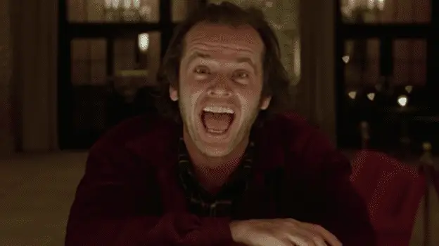 Jack Nicholson as Jack Torrance in The Shining, visible from the chest up, sitting with one arm crossed in front of him, looking directly into the camera with wide eyes, mouth agape in a crazy smile, and eyebrows arched