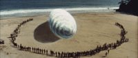 A gaint egg sits in the middle of a beach with hundreds of bystanders staring at it.