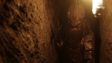 a man in armor walks through a narrow cave, a sword in one hand and a torch in the other
