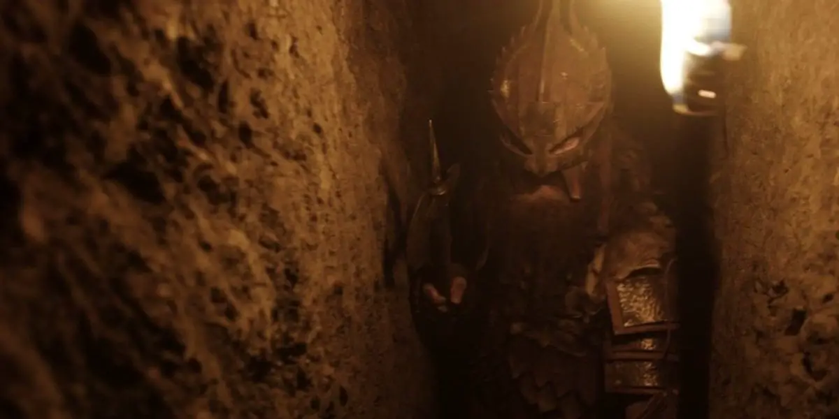 a man in armor walks through a narrow cave, a sword in one hand and a torch in the other