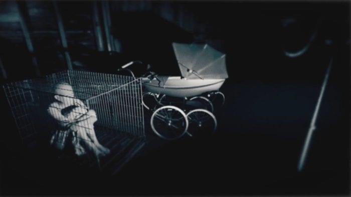 Isabella sits in a cage in a dark room next to a baby carriage while wearing a mask. 