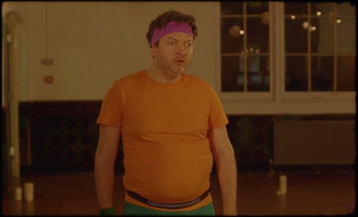 Alex looks befuddled standing in a yoga studio wearing a pink sweatband orange t-shirt and green shorts