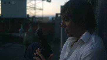 Richard sits on the roof of a building holding a gun