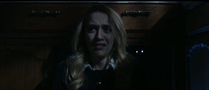 Sasha sits on the floor with her back to an open cabinet where a pair of glowing eyes watches her