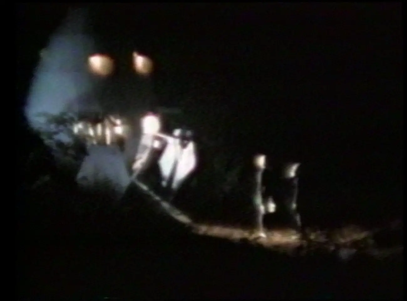 two inhuman figures stand in the distance in the dark in front of a lighted cone shaped ship