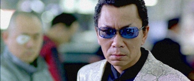 Takashi Miike appearing in Last Life in the Universe wearing blue glasses and a snakeskin print blazer.