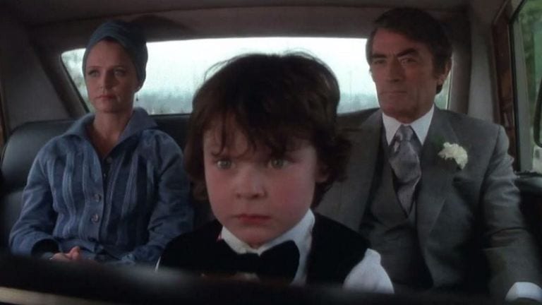 Damien from The Omen sits in front of his parents in the car with a crazed look in his eyes.