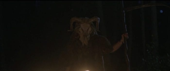 A man wearing pelts and a horned animal skull stands in back of a rising fire