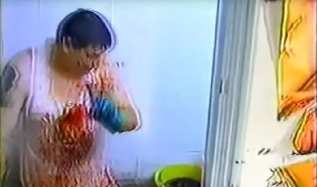 A poor quality image of a white man with dark hair and wearing what looks to be a bloodied white tank top in a white room. He is wearing a blue rubber glove and looking down toward a yellow bucket with some dark colored liquid in it.