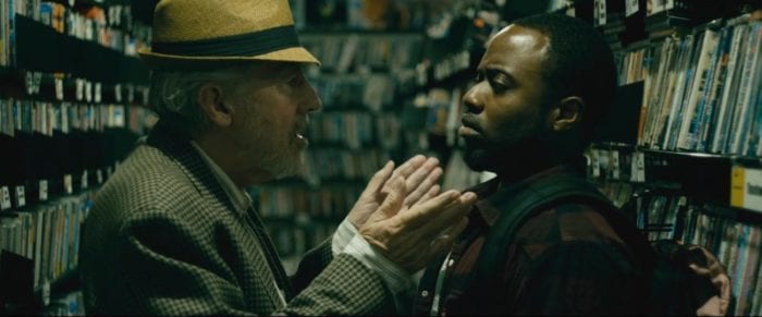 the store owner stands with his hands open in front of Oz, who has backed himself up into one of the video shelves that surround the two men
