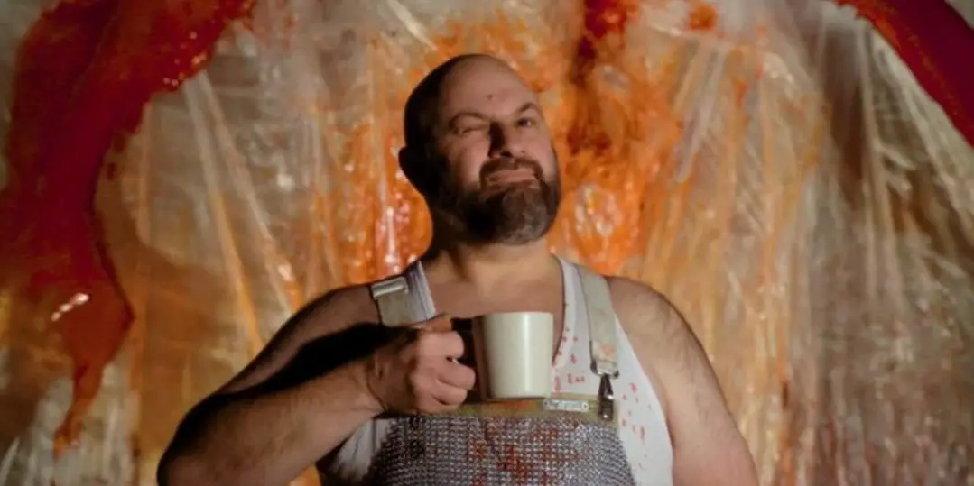 Chester holds his coffee mug and gives a wink at the camera as blood spray hits the plastic sheeting behind him