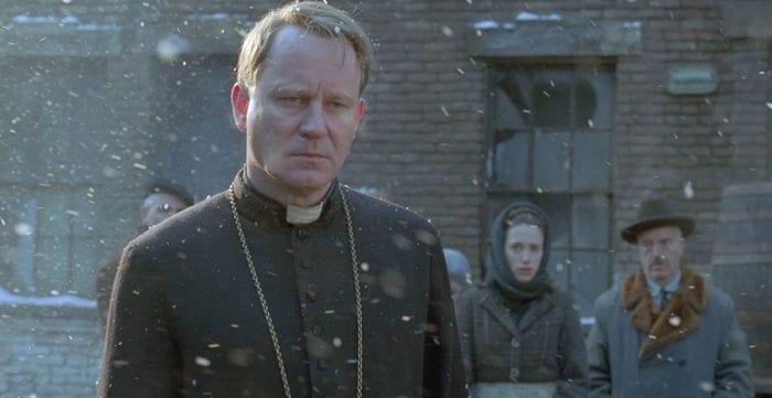  A priest stands in the snow with onlookers behind him.