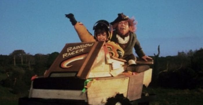A smiling child puts her arm up as she steers a go cart while a green clad leprechaun sits behind her.