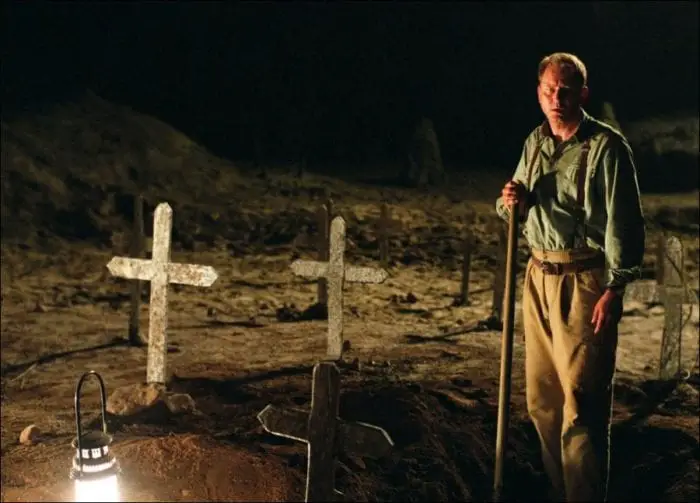 A man stands in a graveyard holding a shovel by lanternlight.