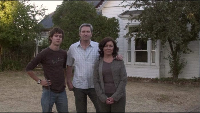 A young man poses with his parents in front of their house. In the dark window behind them a blurry figure can be seen faintly looking on at them.