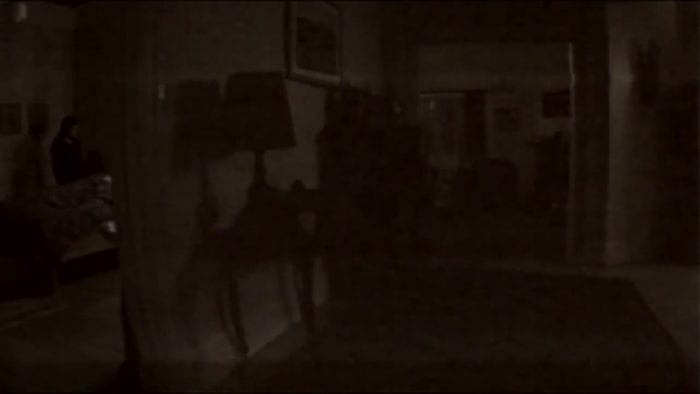 A low quality screenshot of video footage showing a darkened room with a pale, ghostly figure in the corner.