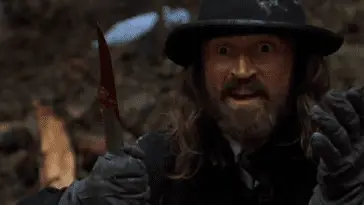 a wide-eyed man in a hat holds a knife covered in blood