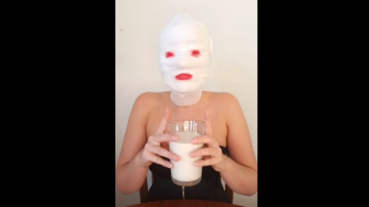 Harley Dee sits at a table with gauze wrapped around her head, lipstick pained on where her eyes and lips would be, raising a glass of milk with both hands