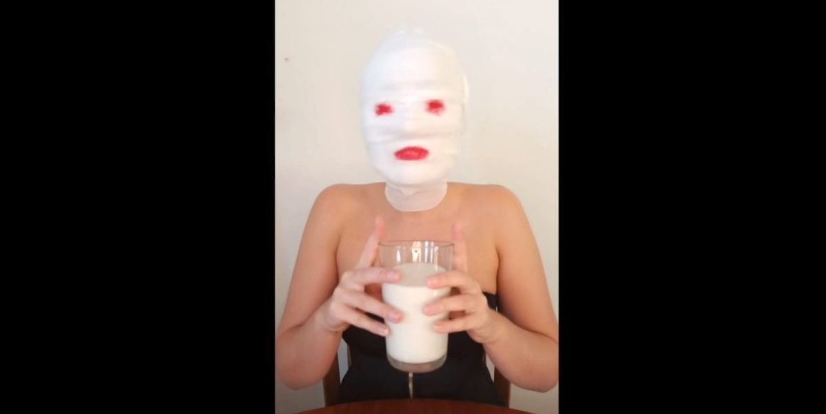Harley Dee sits at a table with gauze wrapped around her head, lipstick pained on where her eyes and lips would be, raising a glass of milk with both hands