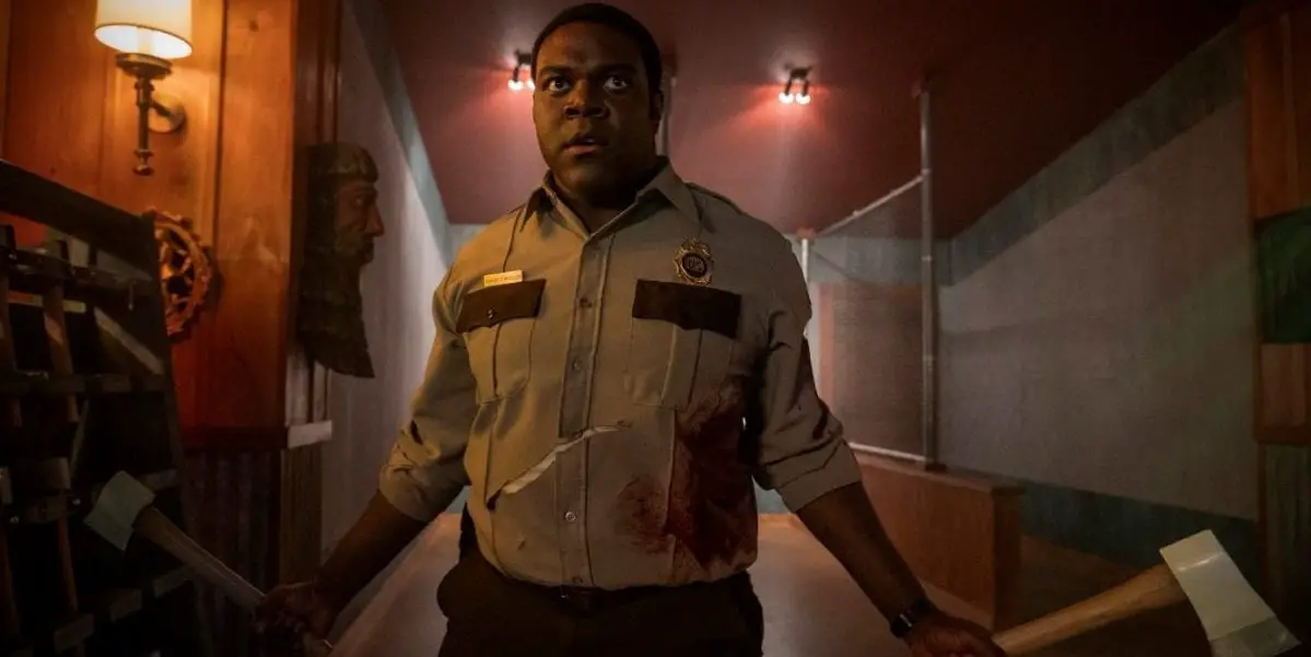 Sam Richardson holds axes in both hands at waist level walking toward the camera down a hotel corridor
