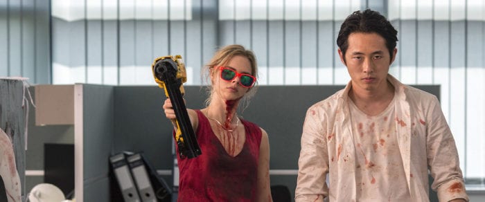 Samara Weaving aims a nail gun while wearing sunglasses. Steven Yuen stands on her left, his clothing flecked with blood