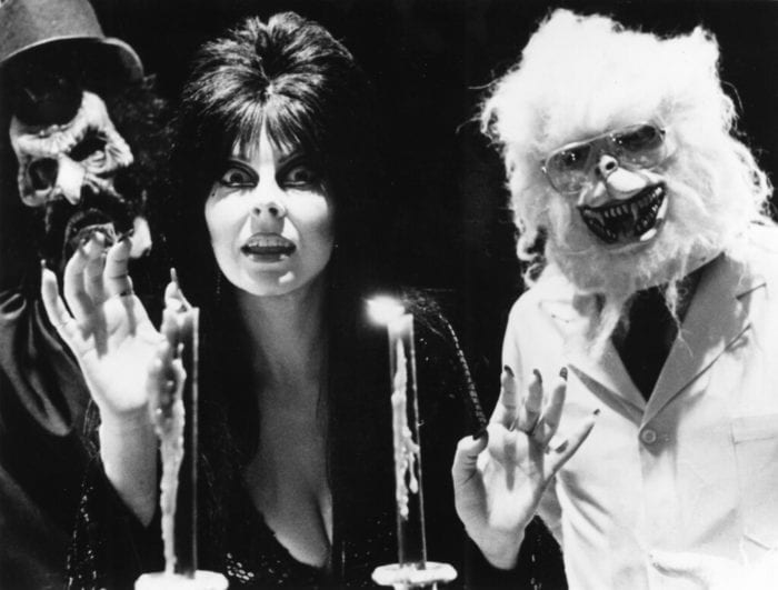 A Halloween style setting with three people all dressed up and waving at the camera. Two candles are lit. In the center is a woman with what looks like a ghoul in a top hat on the left and a white furry werewolf on the right. with a big, dark bouffant hairstyle and elaborate black eye makeup, with a