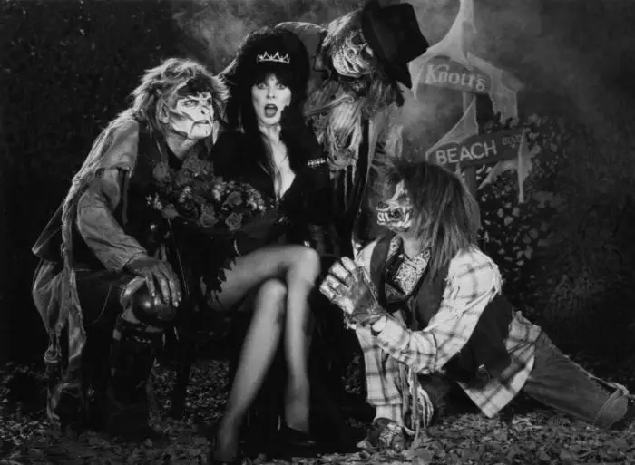 A woman in a black revealing dressed sits in the center of a group of people all dressed up like monsters.