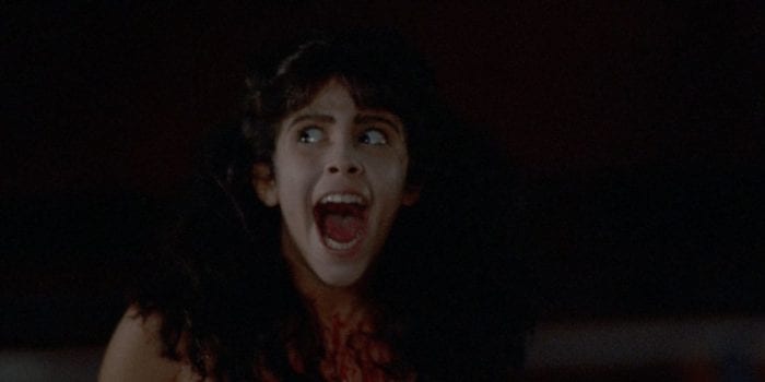 Angela Baker (Felissa Rose) from Sleepaway Camp. She is a pale girl with long, dark hair. Her mouth is wide open in a scream and her eyes are wide. There is blood splattered on her chest.