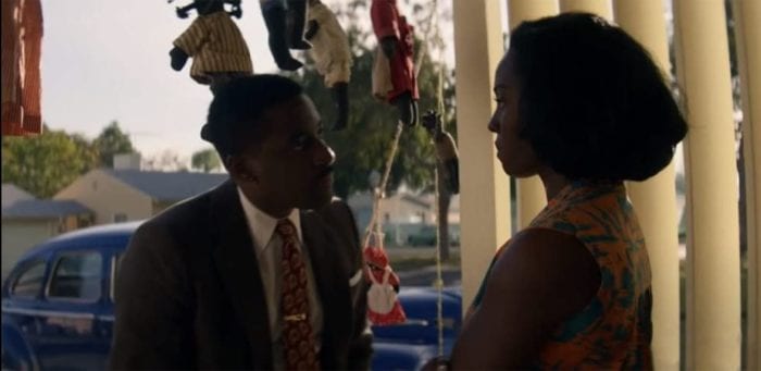 Henry (Ashley Thomas) and his wife, Livia (Deborah Ayorinde), stand on their porch while having a discussion. Numerous pickanniny dolls hang from the roof in the background.