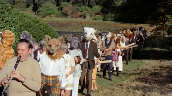 Stretching from the far right background diagonally toward the immediate left foreground , a procession of masked and costumed men and women, some with instruments, others with sticks; visible around them are trees and open fields.