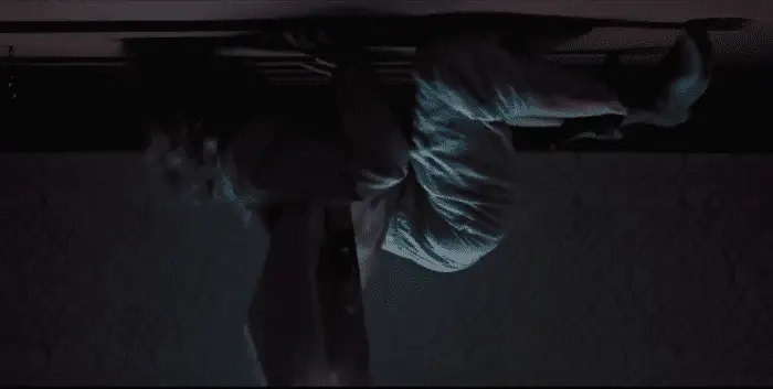 A woman, her face obscured, wearing pajamas and robe, clings on all fours to the ceiling, her hair and robe dangling down.