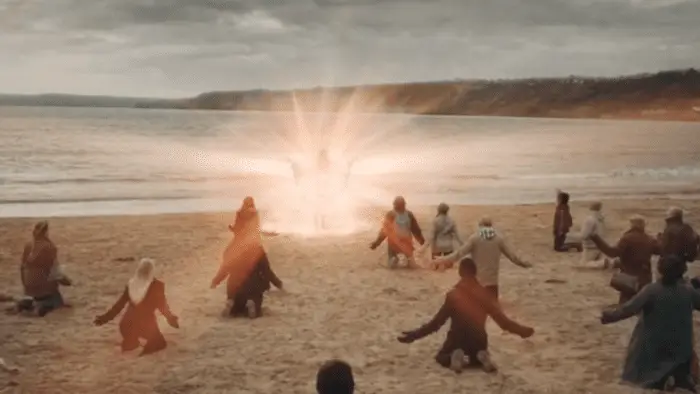 Wide shot of a beach, sand in the foreground, water in the background; on the sand is a crowd of people, seen from behind, kneeling, many with their hands stretched out beside them; in the center of the middle ground is a robed figure, arms uplifted, surrounded by and partially obscured by an intensely bright aura of light.