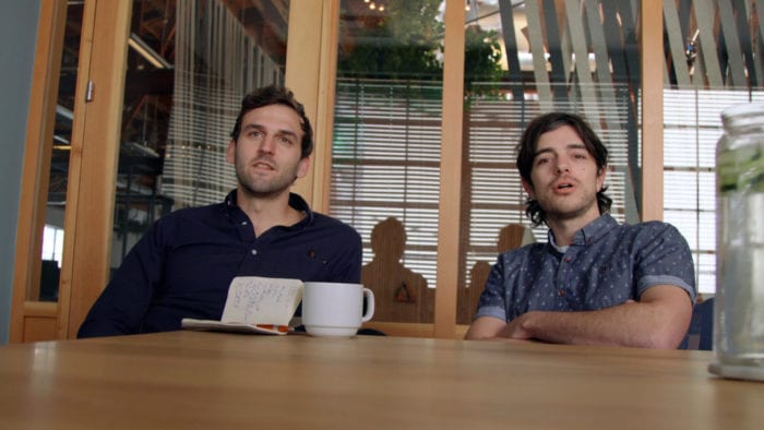 Two men sit at a table and look across at the camera/viewer. A cup of coffee and notebook sit in front of one man on the table.