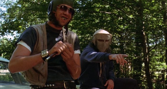 Two men wearing masks, helmets, and sunglasses sit on the hood of a car while one points and the other smiles and picks his nails with a large knife.