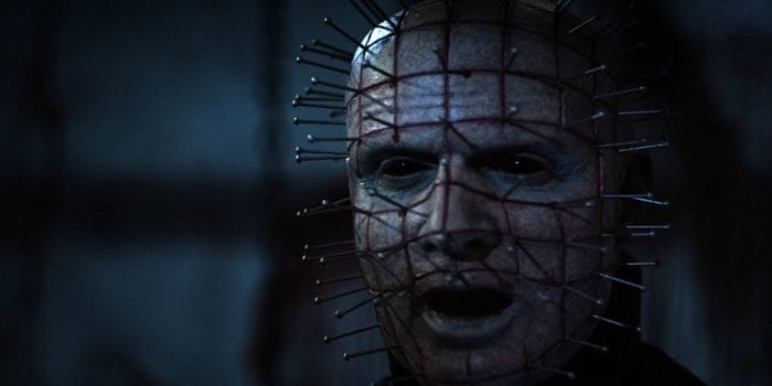 Pinhead (Paul T. Taylor) in a dark room illuminated by dim blueish light. He is a sickly pale with pitch black eyes and graph-like carvings in his face. Nails protrude from his head, each placed neatly and evenly.