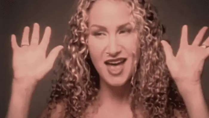 Close-up from the shoulders up of Joan Osborne with long, curly hair and a nose piercing; her hands, one ring on each hand are raised up, palms outward, turned slightly upward.