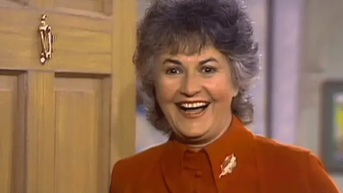 Medium close-up of a woman standing in a doorway, the open door visible to her left; she is wearing a red top with a lapel pin and smiling toward the camera.