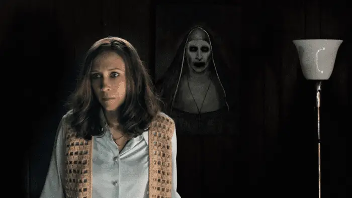 In the foreground just left of center, a Lorraine Warren in white, button-up shirt and an open knit sweater; behind her is blackness, to the far right is a white floor lamp, in the center of the frame, visible behind the woman's left shoulder is a painting of a nun with a pale white face and dark circles around her eyes and mouth.