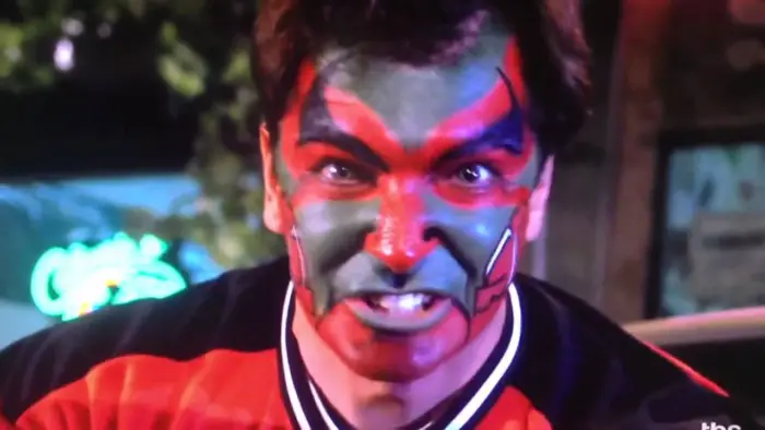 Close-up from the shoulders up of a man wearing a red and black shirt, his face painted red and black; his teeth are bared and he is staring fiercely toward camera in a look of intense anger.