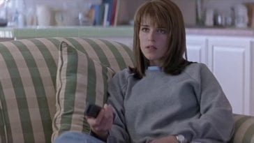 The character Sidney from the Scream franchise, a young woman with shoulder length brown hair, sits on a sofa holding a remote.