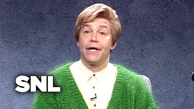 An earnest looking man, facing forward and seen from the chest up sitting in front of a blue background; he is wearing a buttoned up white, collared shirt (with a clip-on microphone visible) and a knit sweater, his hair is brushed up into a high wave in front that comes down just over his right eyebrow.